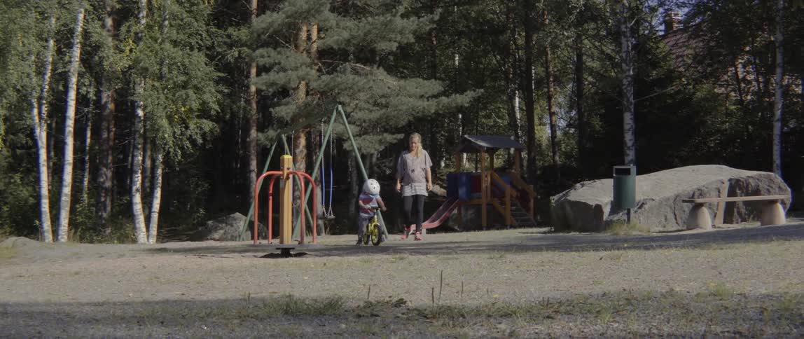 A woman watches as a young toddler boy rides his small bicycle outdoors.