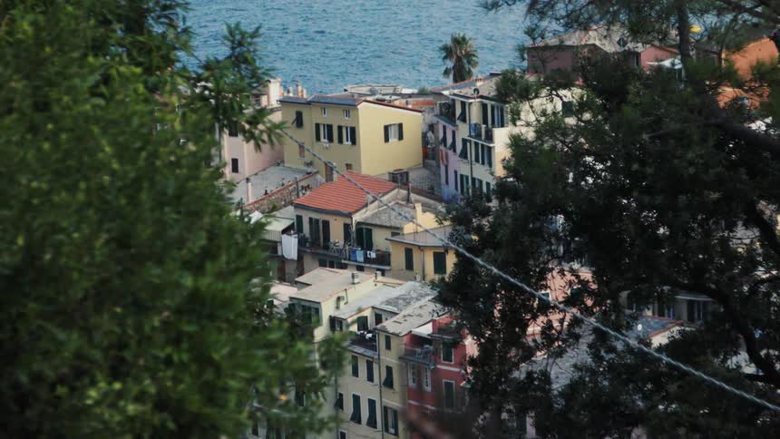 A highangle shot framed by dense green foliage looks down on a cluster of colorful apartment buildings with terra cotta roofs built along the seaside the deep blue water of the ocean is visible just behind the buildings