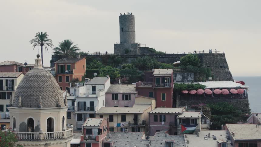 A hillside town with stacked colorful houses in the foreground leads up to a round stone tower and a fortified wall situated on a cliff overlooking the sea