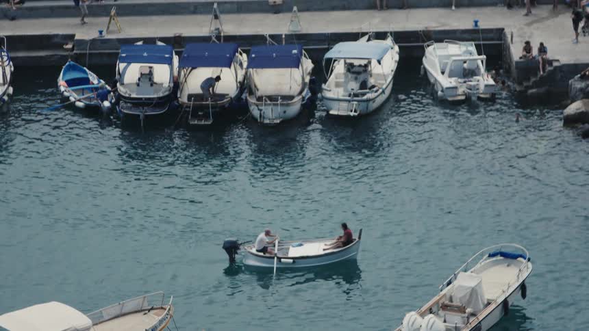 Small boat with two people sailing near a pier with docked vessels