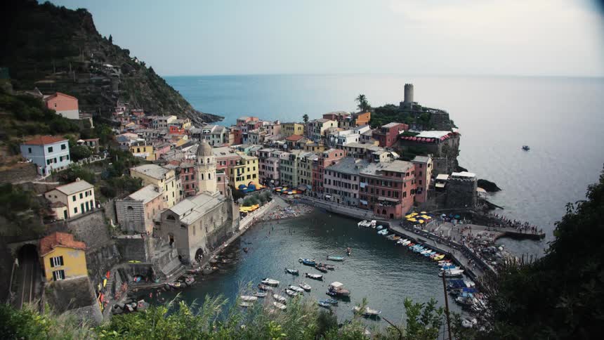 A dense coastal village built around a small natural harbor with moored boats backed by a steep hillside and featuring a round stone tower on a rocky promontory facing the open sea