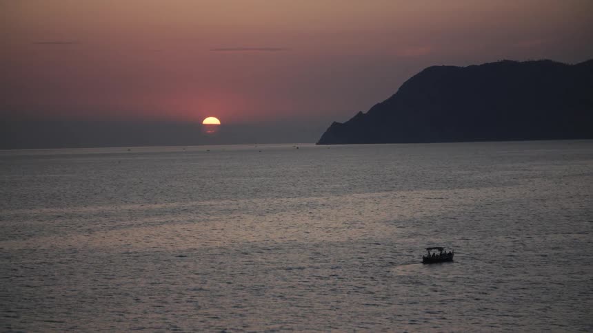 At sunset a boat sails across the ocean with a mountain silhouetted against the glowing orange sun in the background