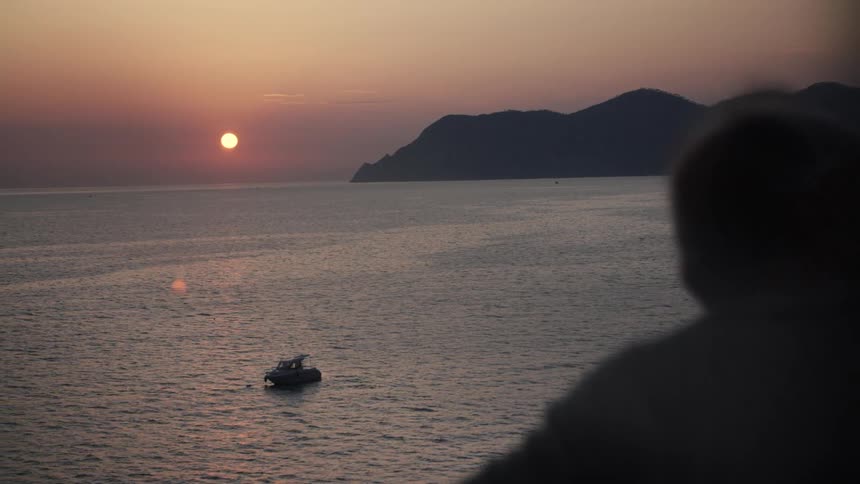 Person watches the sunset over the sea with a boat and mountains in the background