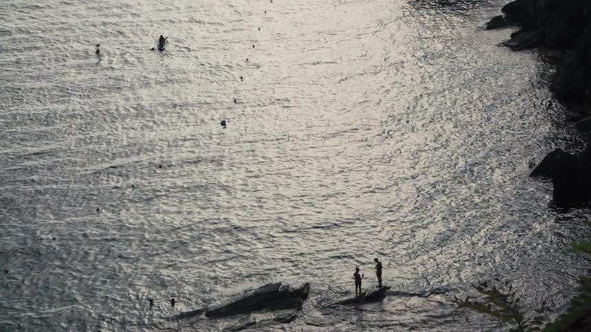 A body of water shimmering with light reflections where two human figures stand on rock outcroppings near the shoreline and a small vessel floats nearby