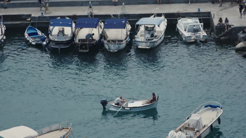 People row a small boat near moored boats at a sunny coastal dock