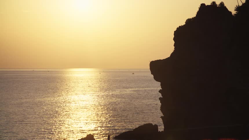 Setting sun reflects on calm sea beside rocky cliff