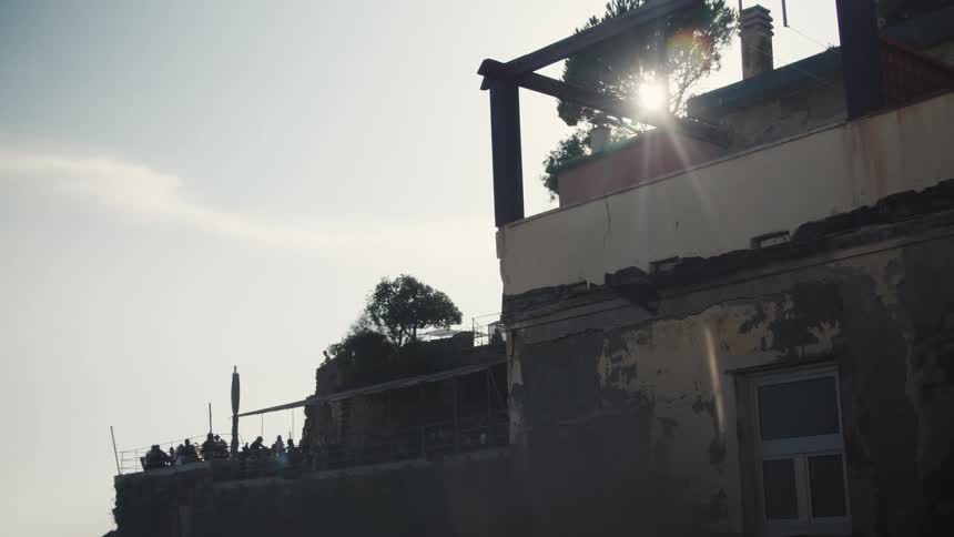 A sunlit outdoor terrace filled with people is partially obscured by the weathered facade of an old building in the foreground