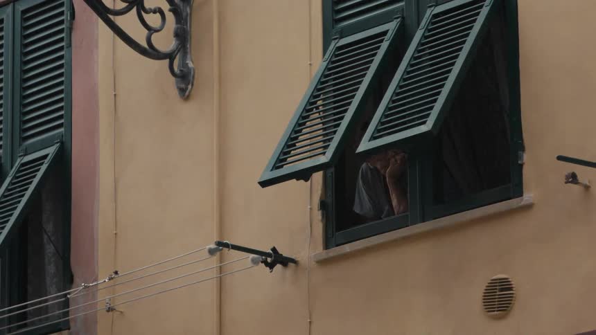 A person looks out of a window with dark green shutters on a peach coloured building while an empty clothesline hangs nearby