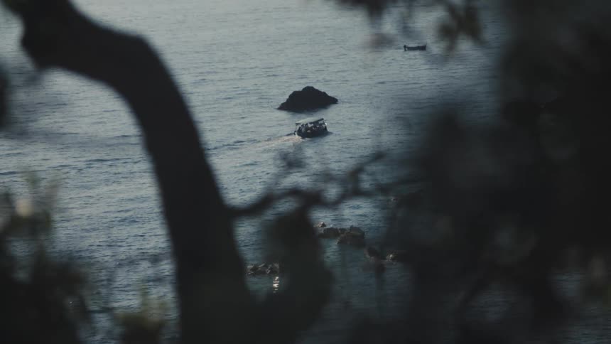 A dark highangle shot is framed by silhouetted tree branches in the foreground creating a natural vignette overlooking the ocean far below a small fishing boat speeds across the rippling dark water near a large rock outcropping