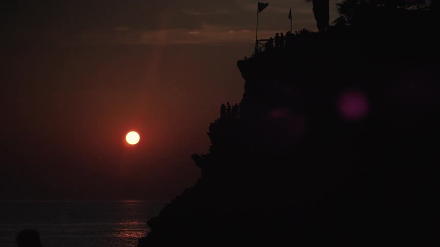 At dusk silhouetted figures crowd a cliffside overlook as the orange sun sinks over a calm sea