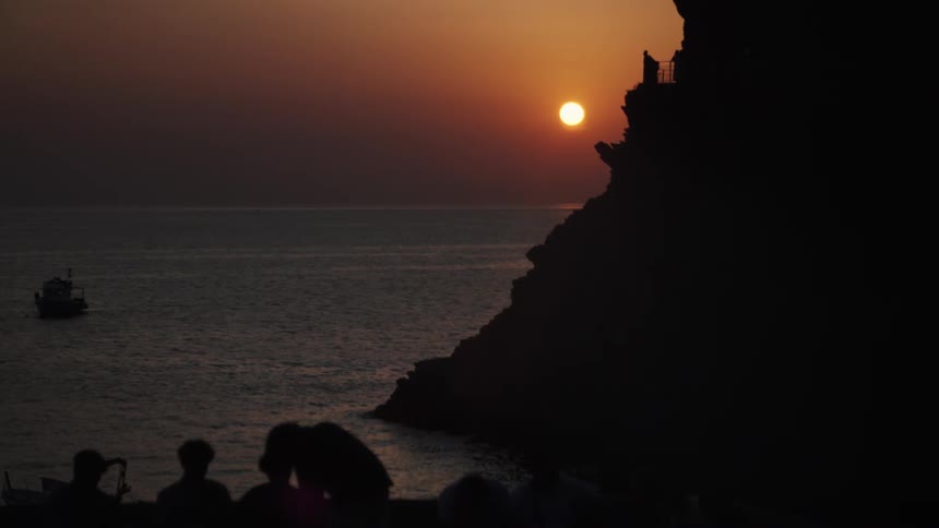 A group of people stands on a rocky shoreline silhouetted against the orange glow of the setting sun in the background