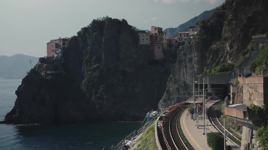 A railway station is nestled into a rugged cliffside along the sea with a quaint village perched above