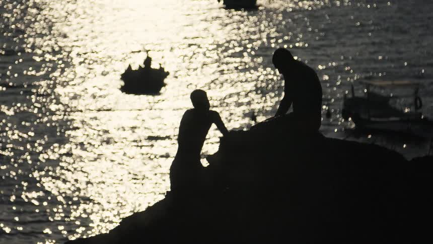 Two people silhouetted on rocks by the sea with boats in the background