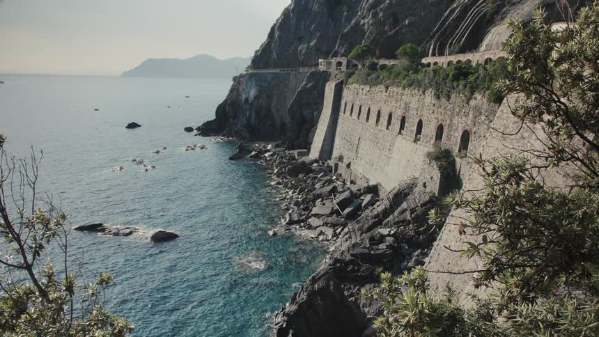 A highangle shot captures a dramatic coastal view featuring a towering cliff face that plunges into the clear deep bluegreen sea a distinctive stone retaining wall with arched openings runs along the base of the cliff supporting a roa