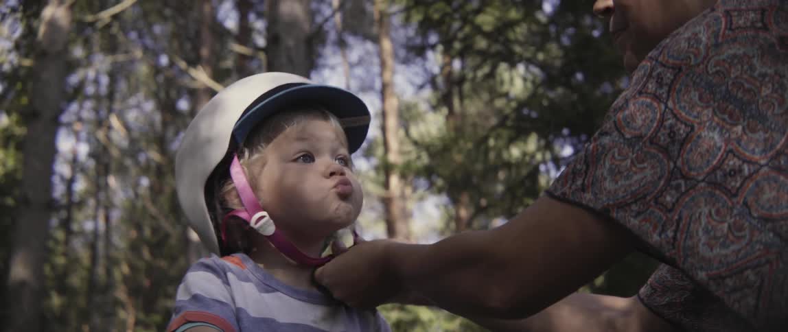 A parent carefully fastens a safety helmet on a young toddler boy outdoors.