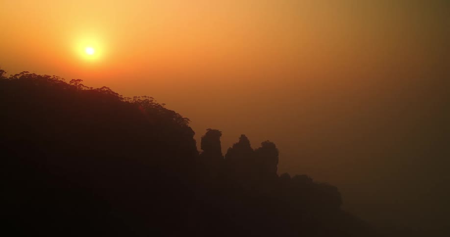 A wide shot captures the silhouette of three prominent rock formations resembling the three sisters of the blue mountains australia against an intense goldenorange sunset or sunrise the sun sits low in the sky casting a bright glow and c