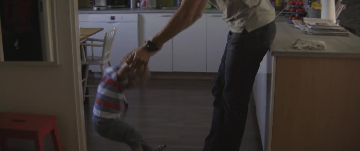 A man playfully twirls a young toddler boy around in a bright indoor kitchen.