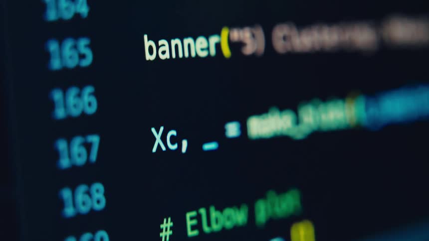 A blurred closeup of a dark computer screen shows numbers beside lines of code with yellow syntax highlights suggesting a developer editor or console in use