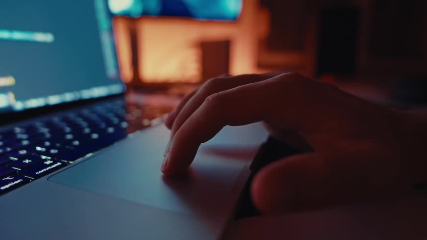 A human hand uses its index finger to manipulate the surface of a laptop trackpad while the screen glows blue and provides ambient light in a darkened room