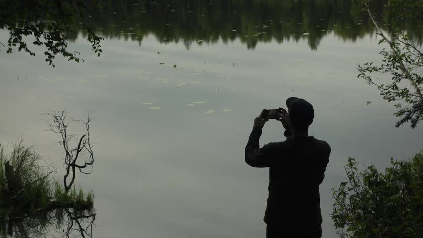 A man by a lake filming the water with a cellphone