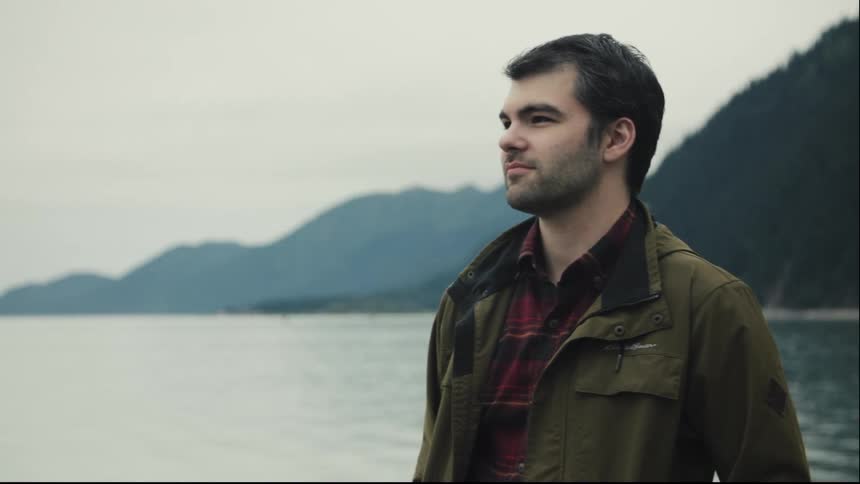 A closeup side profile shot shows a young man with dark hair standing outdoors looking thoughtfully toward a large body of water he is wearing a green jacket over a red plaid shirt with mountains visible in the misty background