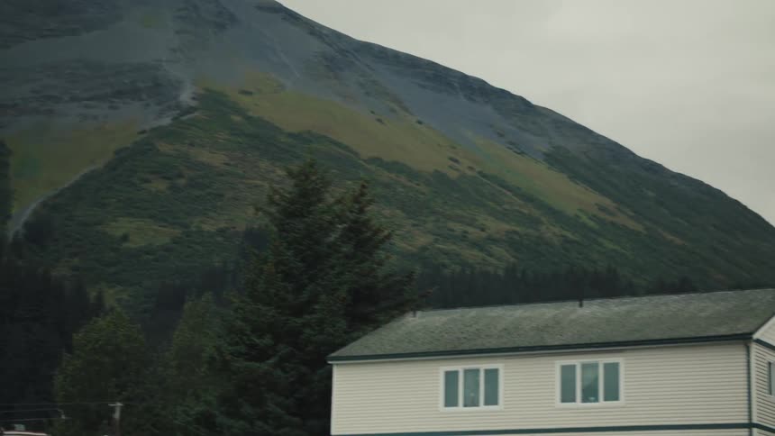 A long shot captures a large conical mountain with a scree slope running down its center the base of the mountain is covered in dark pine trees with a hazy overcast sky in the background
