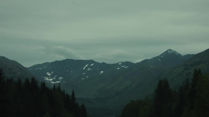 A lowangle long shot captures a dark valley with snowcapped mountains in the background under an overcast sky dark pine trees frame the foreground emphasizing the dramatic and rugged landscape