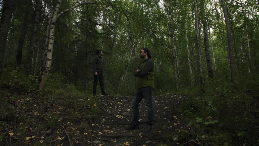 A long shot shows two men standing on a dirt path in a dense dark forest with tall slender trees with one man slightly elevated above the other the overall scene has a cool dim and moody atmosphere due to the lack of direct sunlight penetrating the