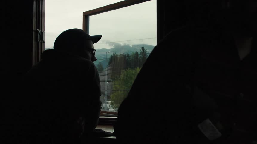 A man rests by the window at midday wearing a hat and glasses as he gazes outside