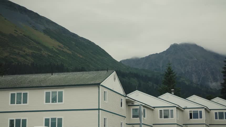 A row of residential buildings stand below misty green mountains under an overcast sky