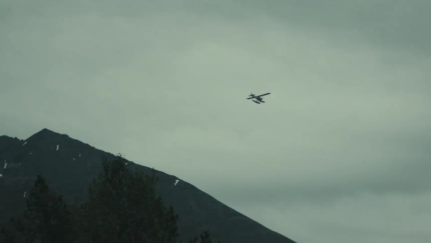 A long shot shows a small black airplane or helicopter flying horizontally in the vast overcast sky centered above a large dark mountain peak the scene is muted and dark giving a sense of vast rugged wilderness