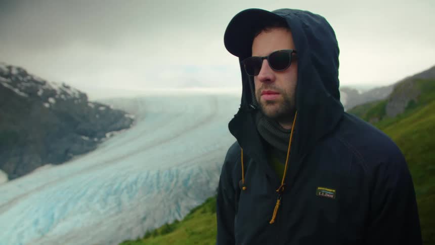 A man wearing a dark jacket and sunglasses stands on a grassy slope with a vast lightblue glacier winding through a deep valley behind him the sky is a misty whitegray creating a cool and rugged atmosphere for the arctic landscape