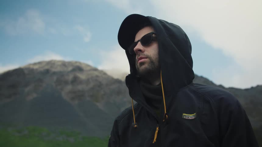 Man in hooded jacket and sunglasses stands outdoors with mountains behind