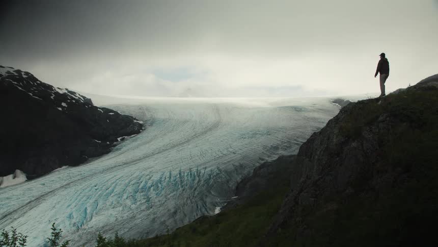 A human silhouette stands on the rocky edge of a steep cliff overlooking a large bluewhite glacier that fills the mountain valley beneath a cloudy bright sky