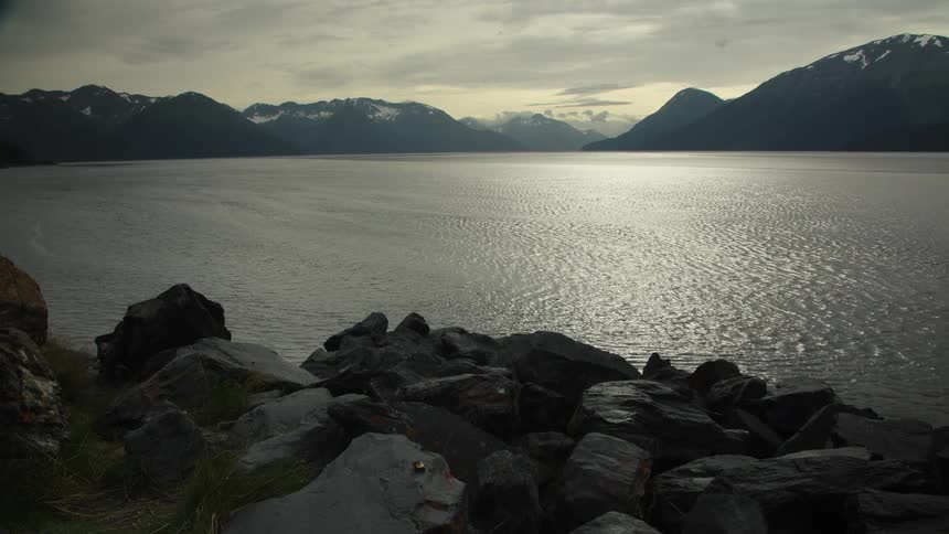Wide shot of rocky shore with sunlit water and snowy mountains at dusk