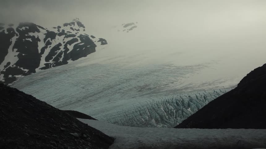 Glacier with visible crevasses nestled between dark rocky slopes and snow under cloudy sky