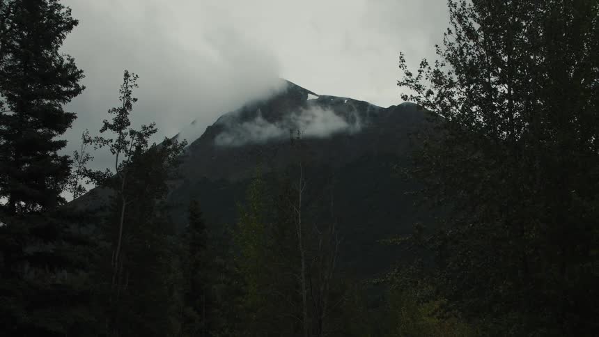 A wide video shows wisps of white fog and low clouds drifting quickly around the dark rocky peak of a steep mountain which is framed by silhouetted evergreen trees against an overcast sky