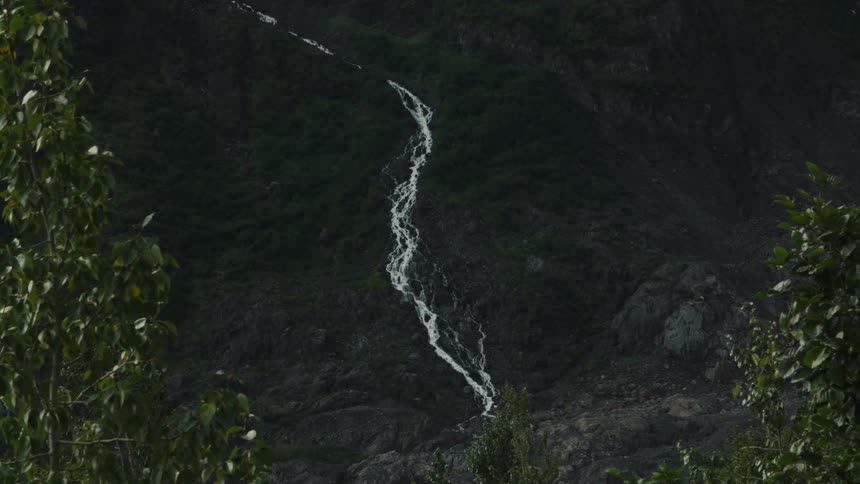 A slender white waterfall snakes its way down a dark rocky mountainside framed by lush green trees