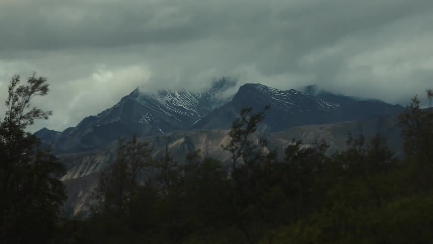 Snowcovered mountains and clouds in the background with dark tree silhouettes in the foreground under dramatic sky