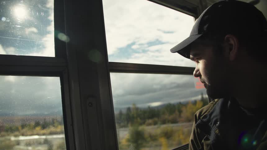Man wearing a baseball cap looking at trees and nature from the window of a moving vehicle during day time