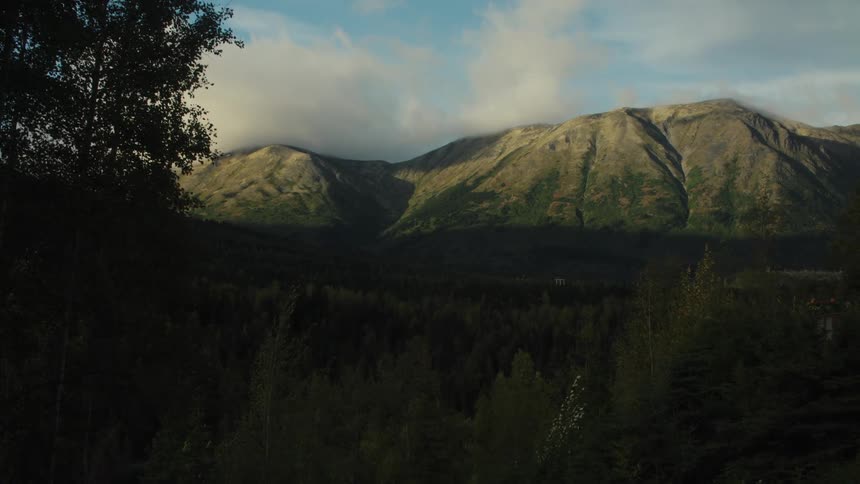 Sunlit mountains with shadowed forested valley partly cloudy sky and foliage framing the scene