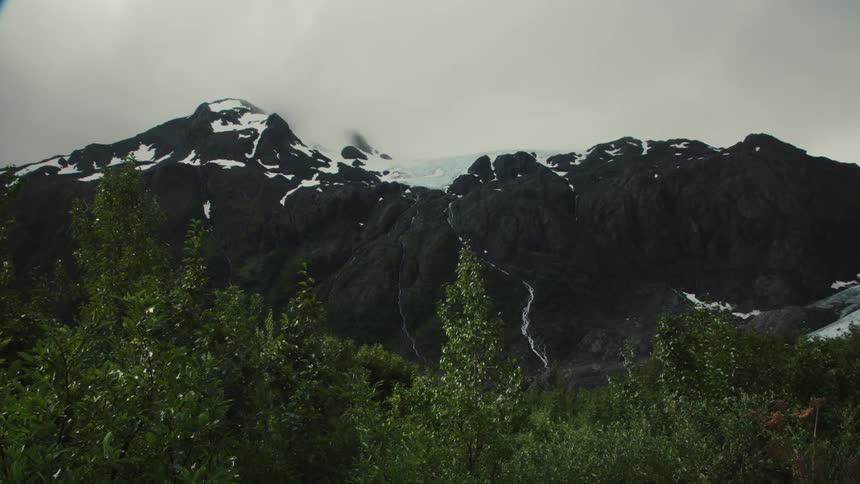 Snow capped mountains rise behind dense green foliage under a cloudy sky