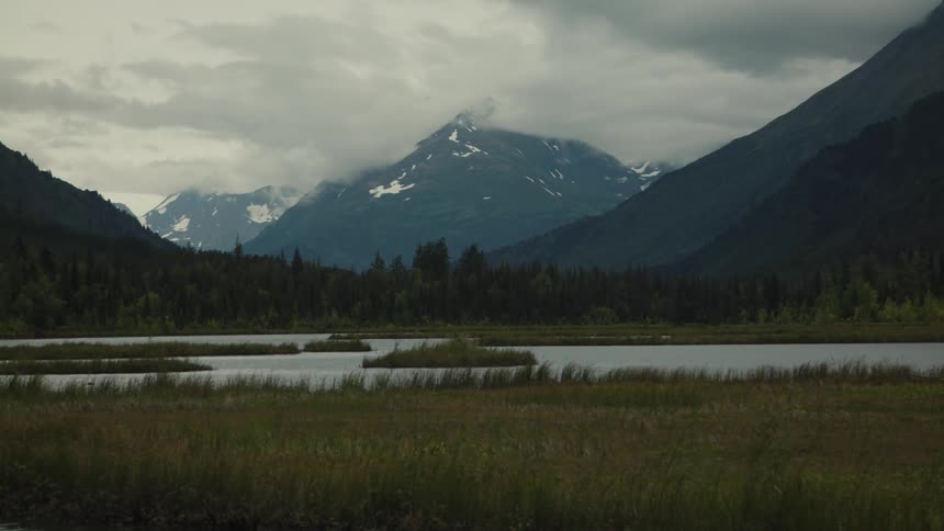 A snow covered mountain range looms over a still lake beneath a cloudy sky