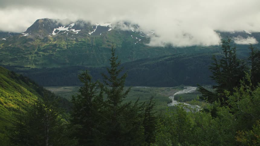 Snowcapped mountains with low clouds surround a lush valley with a winding river
