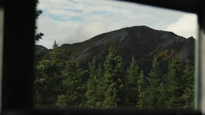 Through the window of a building trees stretch across the wild landscape with a mountainous range rising in the background under the midday sun