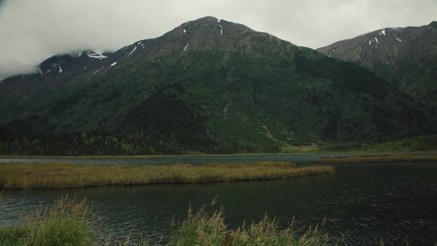 A large mountain peak covered in dark green vegetation rises above a dark lake with a marshy bank in the foreground all beneath a cloudfilled sky