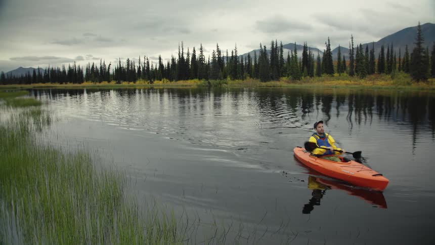 A man in an orange kayak paddles across a still lake flanked by a dense pine forest and distant mountains under a cloudy sky