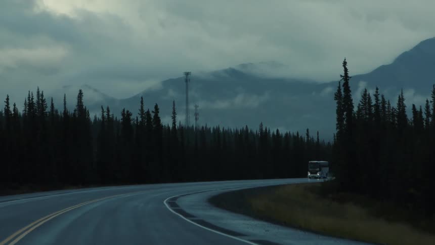 Bus travels along a wet road surrounded by pine trees and mountains shrouded in mist