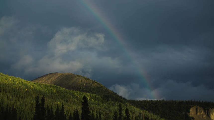 A mountain covered in green vegetation stands under a midday sky with a rainbow arcing overhead