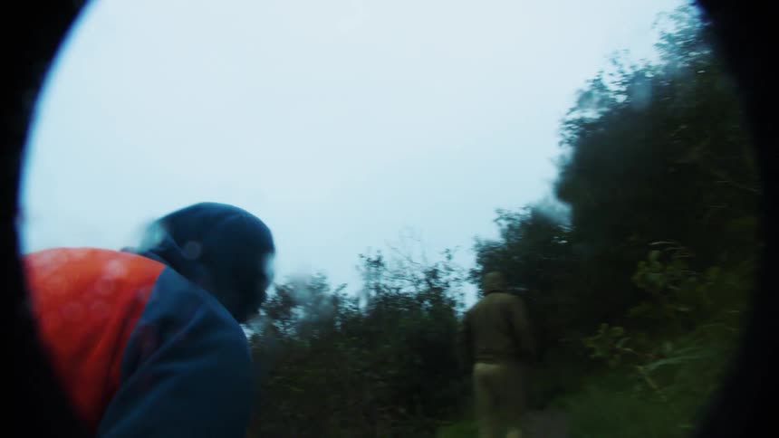 Two people walking along narrow path overgrown with vegetation under cloudy sky